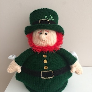 May include: A green knitted teapot cozy shaped like a leprechaun. The cozy has a green hat with a gold buckle, a red beard, and gold buttons.