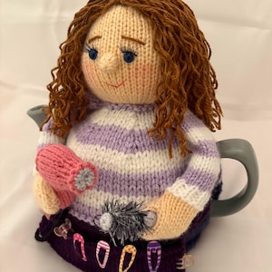 May include: A handmade knitted teapot cozy shaped like a woman with curly brown hair, blue eyes, and a smiling face. She wears a purple and white striped sweater and holds a pink mitten and a black and gray object. The cozy has decorative hair clips.