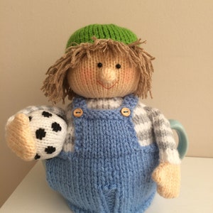 Könnte beinhalten: Eine gestrickte Teekanne in Form einer Person mit grünem Hut, blauer Latzhose und einem Fußball.
