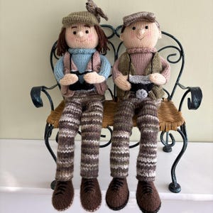 May include: Two handmade knitted dolls, a male and female, are seated on a decorative metal chair. The dolls wear hats, vests, and patterned pants. The female doll wears a blue sweater, and the male doll wears a pink sweater. Both dolls have brown shoes.
