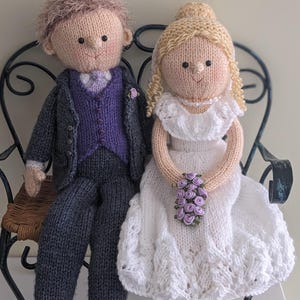 以下が含まれることがあります： 白とグレーの服を着た、ニットの結婚式の人形。紫の花のブーケを持っています。人形は黒い金属製のベンチに座っています。