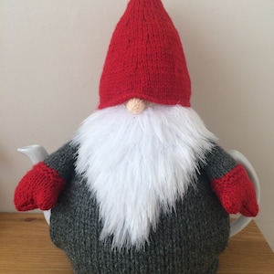 Op de afbeelding: Een gebreide dwerg theepotwarmer met een rode muts, witte baard en grijs lichaam. De dwerg draagt rode wanten.
