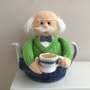 Könnte beinhalten: Eine gestrickte Teekannenhülle in Form einer Person, die einen grünen Pullover, eine blaue Hose und eine Fliege trägt. Die Person hält eine weiße Teetasse mit einem blauen Streifen.