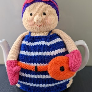 Peut inclure: Une housse de théière en tricot en forme de nageur portant un maillot de bain rayé bleu et blanc, des lunettes roses et un flotteur rose et orange.