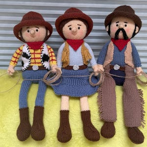 Op de afbeelding: Drie gebreide cowboypoppen, een met een geel en rood vest, een met een blauw vest en een met een bruin vest. Alle drie de poppen dragen bruine cowboyhoeden en bruine laarzen.