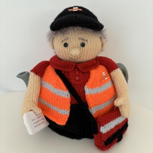 Könnte beinhalten: Gestrickte Teewärmer in Form eines Postboten. Der Wärmer trägt eine schwarze Mütze, ein rotes Hemd und eine orangefarbene Weste mit grauen Reflexstreifen. Er hat eine schwarze Umhängetasche und ein kleines Paket mit dem Text "Mrs. Cosypot Cottage Woolacombe."
