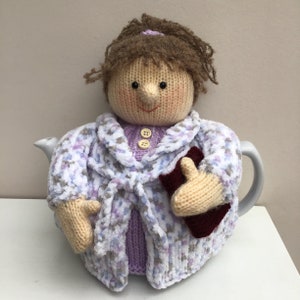 Puede incluir: Una funda de tetera de punto con forma de persona que lleva una bata de color morado y blanco y sostiene un libro. La funda está hecha con un hilo suave y esponjoso.