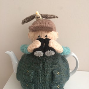 Puede incluir: Una funda de tetera de punto con forma de persona que lleva una gorra marrón, un jersey azul y sostiene unos prismáticos. Un pequeño pájaro está posado en la parte superior de la gorra.