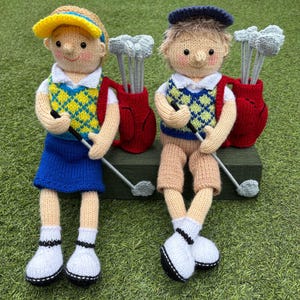 May include: Two knitted dolls dressed in golf attire, each wearing a patterned sweater vest and holding a golf club. One doll wears a yellow visor and blue skirt, while the other wears a navy cap and tan shorts. Both have red golf bags.