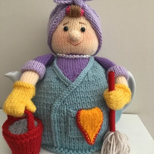 Puede incluir: Una funda de tetera de punto con forma de mujer que sostiene un cubo y una fregona. La mujer lleva un vestido morado, una diadema morada y guantes amarillos. La funda de la tetera tiene un parche en forma de corazón en la parte delantera.
