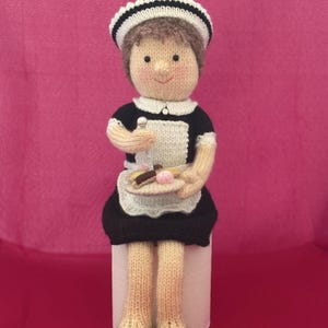 Op de afbeelding: Een gebreide pop gekleed in een zwart-wit dienstmeisjesuniform met een bijpassend kapje. De pop heeft bruin haar, een perzikkleurig gezicht en houdt een klein bordje vast met miniatuurvoedsel, waaronder een roze bolletje.