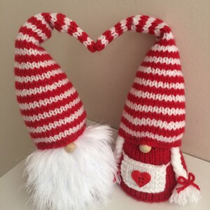 May include: Two knitted gnomes in red and white striped hats, forming a heart shape with their heads. The gnomes have white beards and are wearing red and white outfits. One gnome has a red heart on its apron.