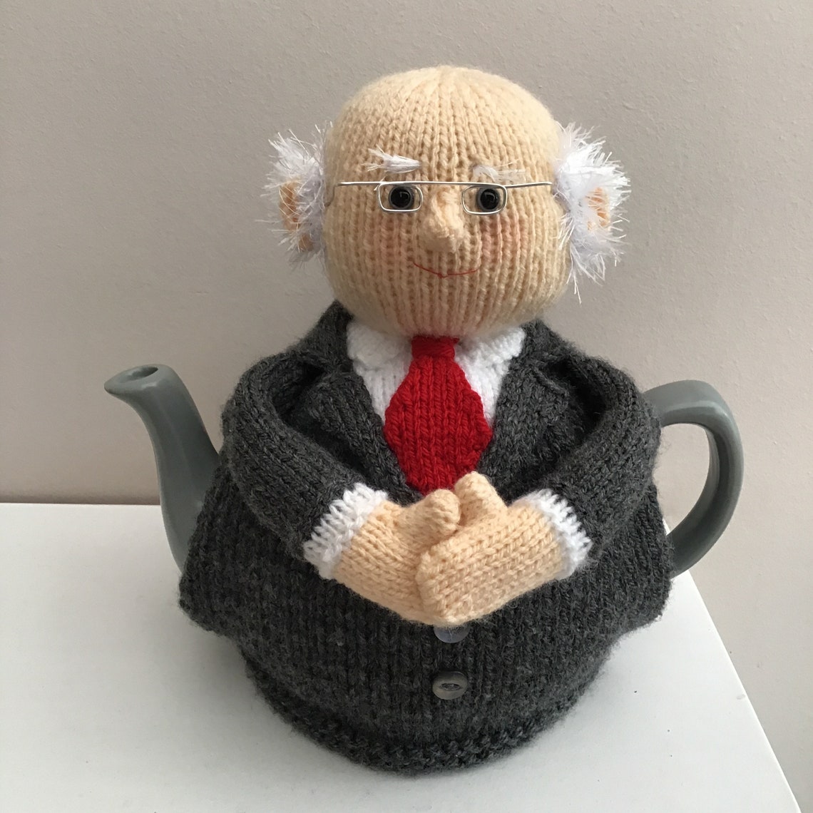 May include: A knitted teapot cosy in the shape of a person wearing a grey suit, a red tie, and glasses.