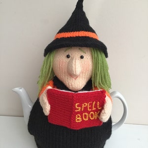 May include: A black knitted teapot cozy shaped like a witch with a green wig, orange and black pointed hat, and holding a red book that says "SPELL BOOK" in yellow letters.