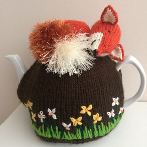 May include: A brown knitted teapot cosy with a fox design. The fox is orange and white and has a fluffy tail. The cosy has embroidered daffodils around the bottom.