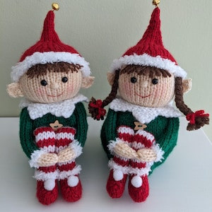 Puede incluir: Dos muñecas elfo de punto con gorros rojos, jerséis verdes y calcetines a rayas blancas y rojas. Los elfos están sentados con las piernas cruzadas y las manos juntas en sus regazos.