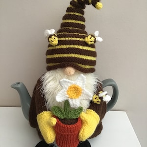 May include: A brown and yellow striped knitted teapot cosy with a bee hat and a flower pot in its hands. The cosy is sitting on a white surface.