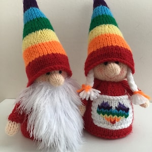 Puede incluir: Dos adornos de gnomo de punto de arcoíris. Un gnomo tiene barba blanca y el otro tiene un corazón de arcoíris en su delantal.