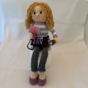 May include: A handmade knitted doll of a hairstylist. The doll has blonde curly hair, a striped purple and white top, grey trousers, and purple shoes. It holds a pink hairdryer and a small brush, with scissors and clips on its apron.