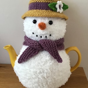 May include: A white knitted teapot cozy shaped like a snowman with a purple scarf, a yellow hat with a purple band and a white flower, and a yellow teapot.