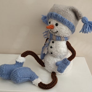 Könnte beinhalten: Ein gestricktes Schneemann-Spielzeug mit einem braunen, weißen und blauen Hut, einem blauen Schal und blauen Fäustlingen. Der Schneemann sitzt auf einer weißen Oberfläche.