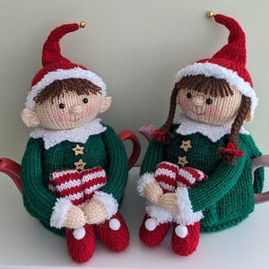 Puede incluir: Dos adornos de calienta-tetera de elfo tejidos en verde, rojo y blanco. Los elfos llevan gorros rojos, jerséis verdes y botas rojas con ribetes blancos. Están sosteniendo calcetines a rayas rojas y blancas.