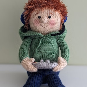 May include: A knitted toy person wearing a green hoodie and blue jeans. The toy has red hair and is holding a grey video game controller.