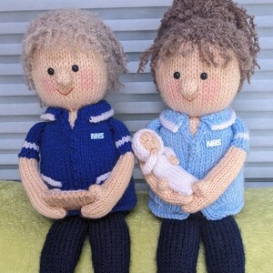 May include: Two knitted dolls dressed as nurses, one holding a smaller knitted doll. The dolls are wearing blue and light blue uniforms with white trim and NHS badges.