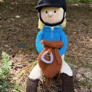 May include: A knitted equestrian doll wearing a black riding hat, blue jacket, and brown riding boots. The doll has blonde hair and a brown saddlebag. The doll is sitting on a bed of leaves and twigs.