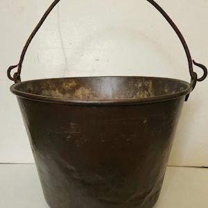 May include: A vintage, dark brown metal bucket with a curved, rusty handle. The bucket has a slightly tapered shape and shows signs of age and wear, suggesting it is an antique or a well-used item. The handle is attached with simple hooks.