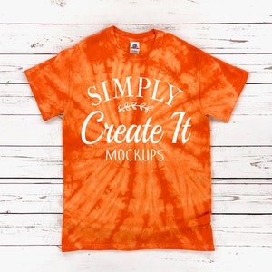 May include: An orange tie-dye t-shirt featuring white text that reads SIMPLY, Create It, and MOCKUPS, with a small decorative leaf branch graphic centred between the first two lines of text. The shirt has short sleeves and a crew neckline.