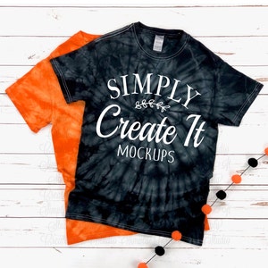 May include: A black tie-dye t-shirt layered over an orange tie-dye t-shirt. The black shirt features white text that reads SIMPLY Create It MOCKUPS with a small leaf graphic. The shirts have short sleeves and a crew neckline with a visible neck tag.