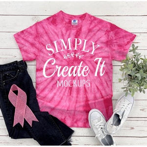 May include: A bright pink tie-dye t-shirt featuring the white text "SIMPLY Create It MOCKUPS" with a small leaf graphic. The shirt is displayed alongside black denim jeans, a light pink ribbon, a small green potted plant, and white sneakers.