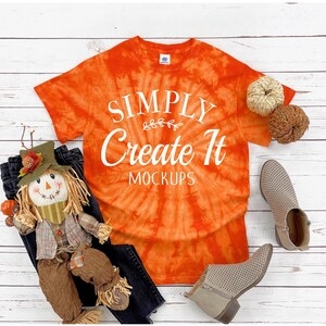 May include: An orange tie-dye t-shirt with white text that reads SIMPLY Create It MOCKUPS, accompanied by a scarecrow doll, two small woven pumpkins, and a pair of beige perforated ankle boots with side zippers on a white wooden surface.