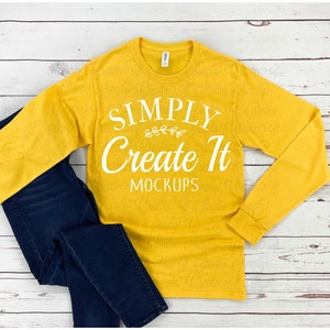 May include: A mustard yellow long-sleeve t-shirt with the text "SIMPLY Create It MOCKUPS" in white. The shirt is laying flat on a white wooden surface with a pair of blue jeans.