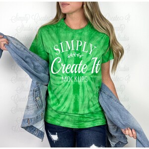 Puede incluir: Una camiseta verde brillante con estampado tie-dye que presenta el texto blanco "SIMPLY Create It MOCKUPS" y un pequeño gráfico de hojas sobre la palabra "Create". La camiseta se lleva con una chaqueta vaquera azul y vaqueros oscuros.