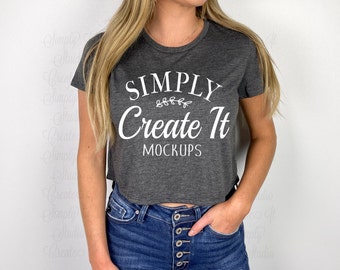 May include: A gray cropped t-shirt with short sleeves featuring the text "SIMPLY Create It MOCKUPS" in white. The shirt is being worn by a person with long blonde hair and blue jeans.