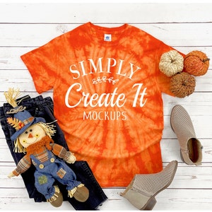 May include: An orange tie-dye t-shirt with white text that reads SIMPLY Create It MOCKUPS, accompanied by a scarecrow doll, dark denim jeans, two small decorative pumpkins, and a pair of beige perforated ankle boots with side zippers.