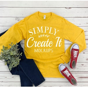 May include: A mustard yellow long-sleeved t-shirt with the text "SIMPLY Create It MOCKUPS" in white. The shirt is laid flat on a white wood background with a pair of red canvas shoes and blue jeans.