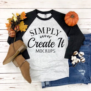 May include: A heather gray and black raglan baseball shirt with the black text "SIMPLY Create It MOCKUPS" and a small leaf graphic. The shirt is surrounded by two brown suede ankle boots, a small orange pumpkin, and cotton stems.