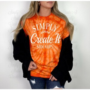 May include: An orange tie-dye t-shirt featuring the white text Simply Create It MOCKUPS with a small leaf graphic. The shirt is worn under a black long-sleeve layer with dark blue distressed denim jeans, creating a casual layered look.