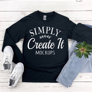 May include: Black long-sleeved t-shirt with white text that reads "SIMPLY Create It MOCKUPS" with a small leafy design above the word "Create". The shirt is laid flat on a white wood surface with a pair of white trainers and blue jeans.