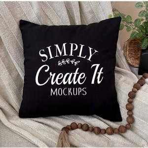 Puede incluir: Cojín negro con texto blanco que dice "SIMPLY Create It MOCKUPS". El cojín está sobre una superficie blanca texturizada con una guirnalda de cuentas de madera marrón.
