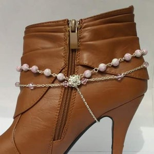 May include: A brown leather high-heeled boot with a decorative silver chain anklet. The anklet features pink and white beads and a central floral design with a dangling chain. The boot has a zipper closure.