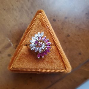 May include: A gold ring with a cluster of pink and white gemstones set in a flower shape. The ring is displayed in a triangular velvet box.