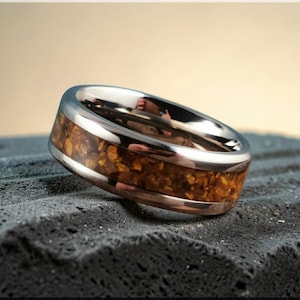 May include: A silver ring with a band of amber-coloured, textured material. The ring is set on a dark, textured surface. The ring's design features a polished, metallic finish.