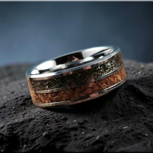 May include: A silver ring with a distinctive design. The ring has a polished silver band, a central section of dark, speckled material, and a band of light brown wood. The ring is set against a dark, textured background.