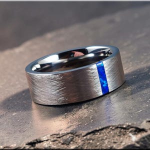 May include: A brushed silver ring with a vertical inlay of blue stone. The ring has a wide band and a textured surface. The blue stone inlay adds a pop of color to the otherwise monochromatic design. A modern piece of jewelry.