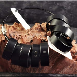 May include: A set of black ring sizers, each with a different size number, connected by a black wire. The ring sizers are displayed on a piece of wood with a light-colored, angled support.