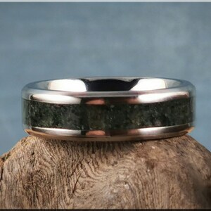 K&ouml;nnte beinhalten: Ein silberner Ring mit breitem Band und einem dunkelgr&uuml;nen, strukturierten Einsatz. Das polierte Metall des Rings kontrastiert mit dem nat&uuml;rlichen, steinartigen Aussehen des Einsatzes. Der Ring ist vor einem neutralen Hintergrund ausgestellt.